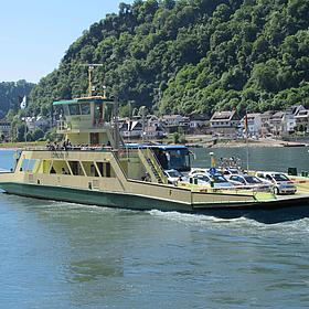 It shows a car ferry on a river. In the background, there is a hill with a forest.