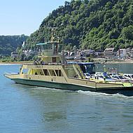 It shows a car ferry on a river. In the background, there is a hill with a forest.