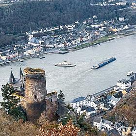 From a castle on a hill, one looks into the Rhine Valley, with two ships and a village visible.