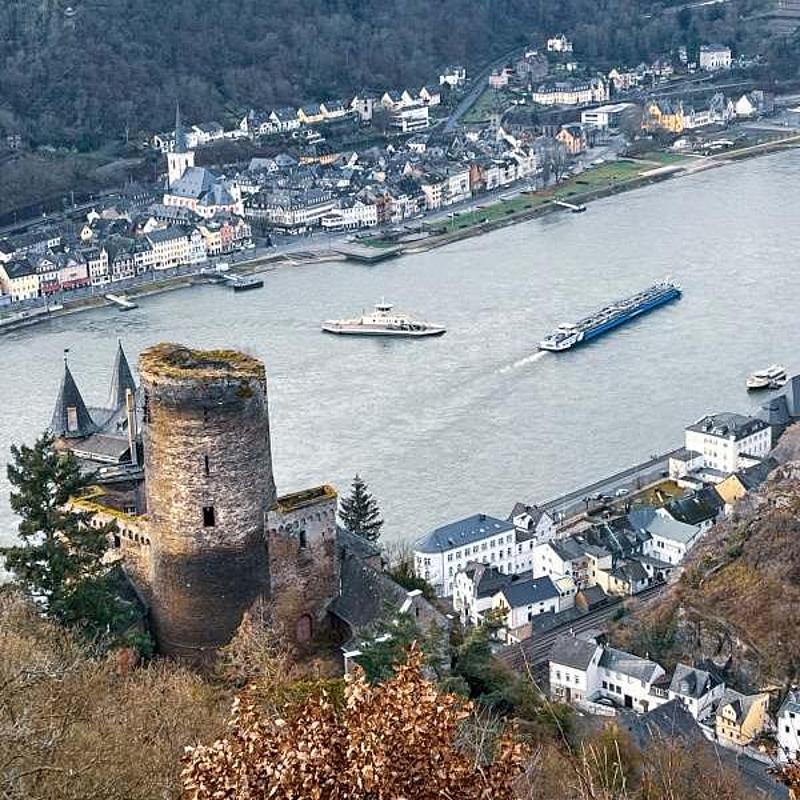 From a castle on a hill, one looks into the Rhine Valley, with two ships and a village visible.