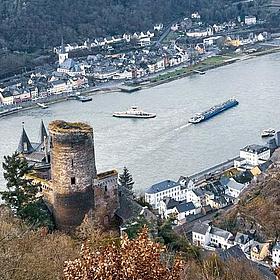 Von einer Burg auf einem Hügel blickt man ins Rheintal, mit zwei sichtbaren Schiffen und einem Dorf.