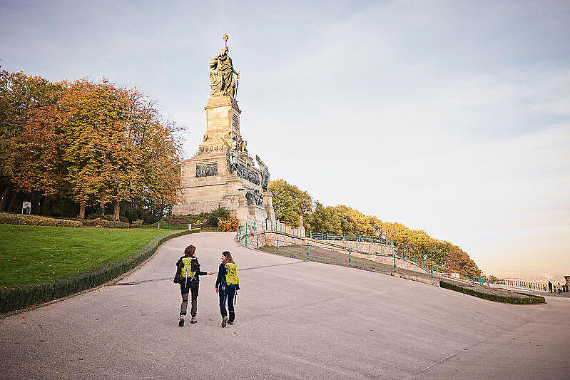 Zwei Wanderinnen sind von hinten zu sehen, wie sie auf ein großes Denkmal zugehen.